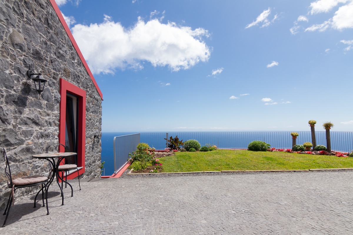 A stone exterior of the house is shown, featuring red trim that contrasts against the grey walls. A small bistro table with two chairs is positioned on the stone patio. Lush green grass and flower beds, along with a railing, overlook a vast view of the Atlantic Ocean under a clear blue sky.