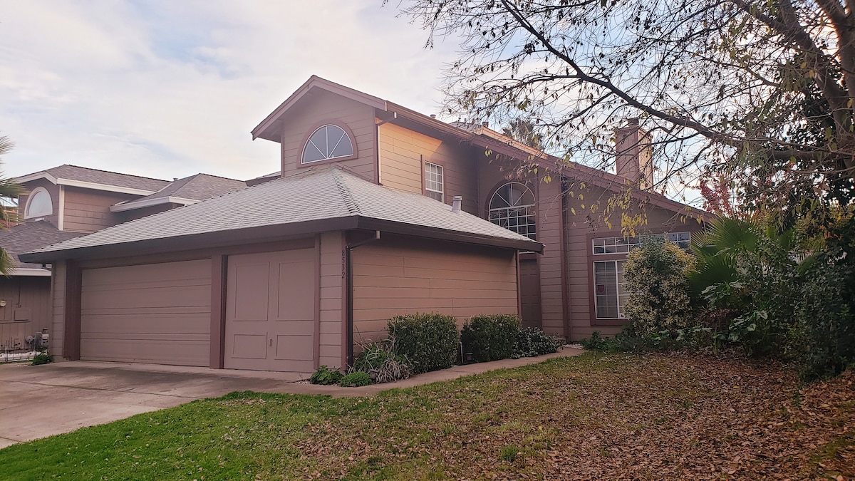 A spacious home is depicted, featuring a two-story structure with a light brown exterior. Large windows allow natural light to fill the interior, while a neatly landscaped front yard includes various shrubs and trees. A driveway leads to the attached garage.