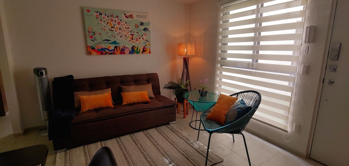 A living area is presented, featuring a brown sofa with colorful accent pillows and a glass coffee table. Soft light emanates from a standing lamp beside the window, which has striped blinds allowing natural light to filter through. A decorative wall artwork adds a vibrant touch.