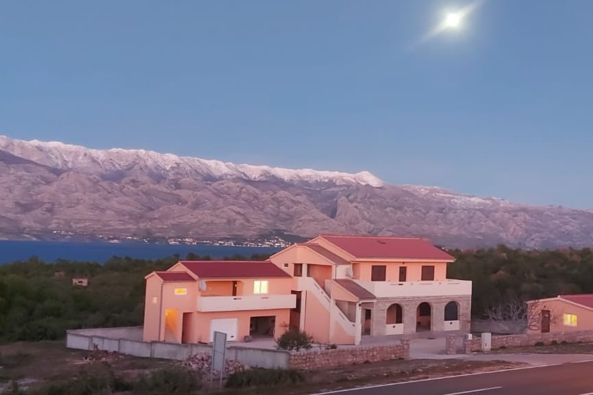 A spacious multi-level building is observed, featuring a red-tiled roof and light-colored walls. Surrounding mountains, capped with snow, serve as a backdrop to the property. The calm waters of a nearby body of water reflect the evening sky, creating a serene atmosphere.