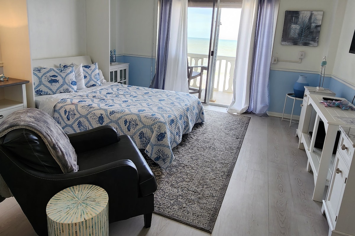 A spacious studio with a king bed features light blue and white bedding. Natural light fills the room from sliding glass doors leading to a private balcony with ocean views. A comfortable chair is positioned nearby, and a flat-screen TV is mounted on the wall.