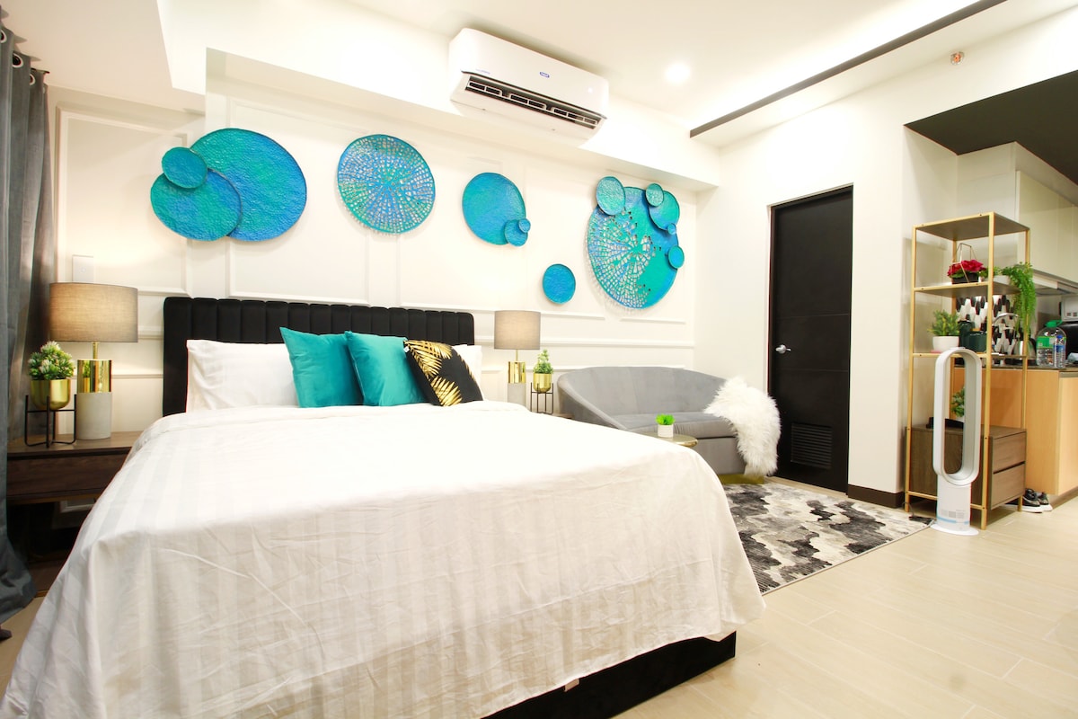 A spacious bedroom features a black upholstered bed topped with a white blanket and decorative teal pillows. Colorful wall decor adds vibrancy, while a cozy gray lounge chair with a white throw complements the modern aesthetic. Soft lighting creates a welcoming atmosphere.
