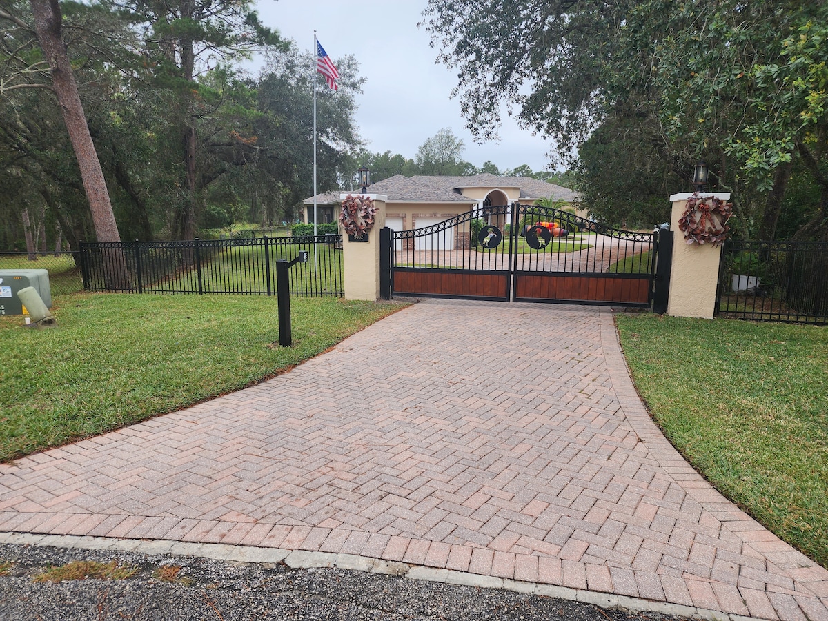 A gated entrance is viewed, featuring a pair of wooden doors adorned with wreaths. The driveway is made of patterned brick, bordered by lush green grass. A flag is displayed prominently, and trees provide a natural setting in the background.