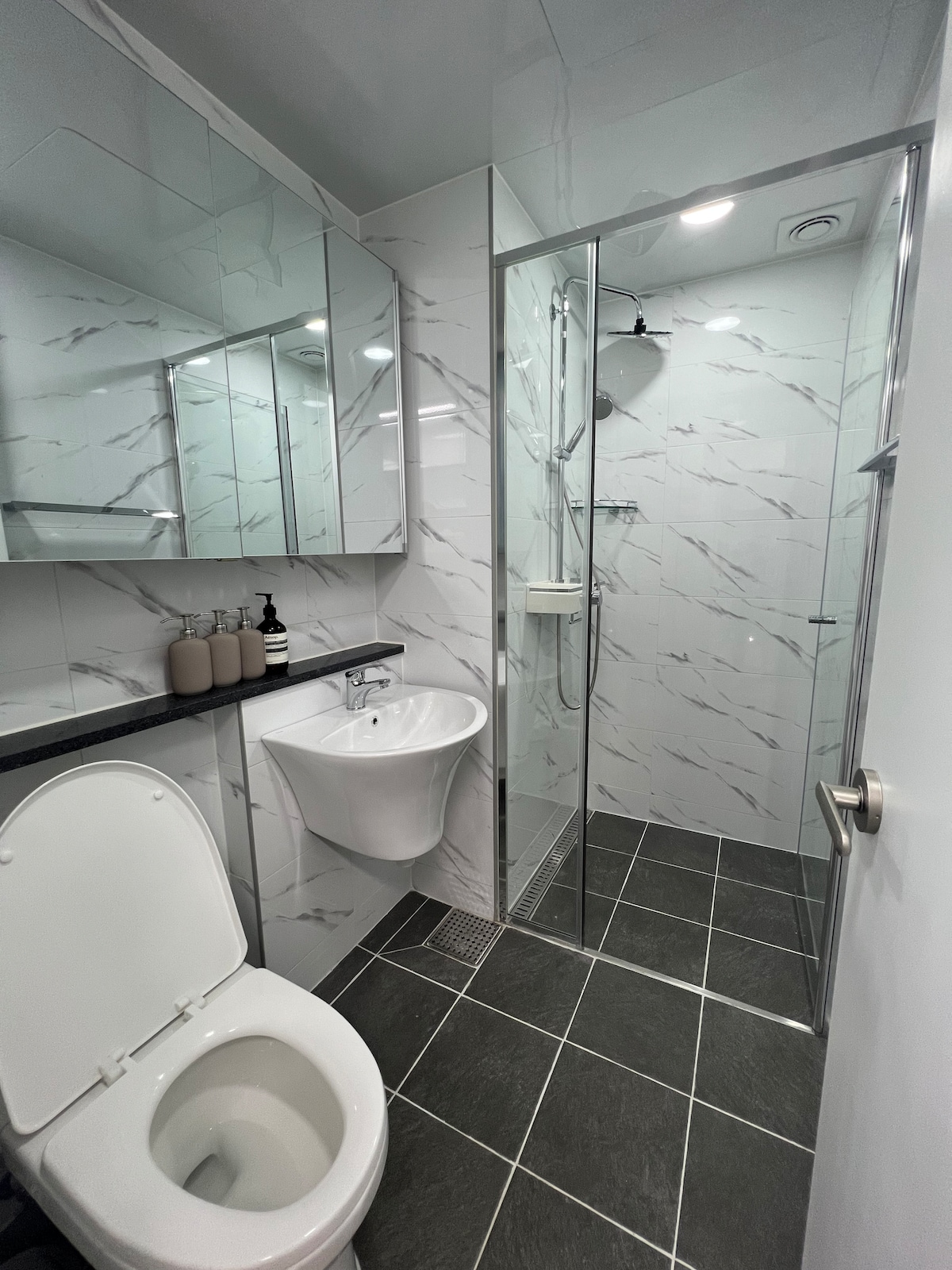 A modern bathroom features sleek marble-patterned walls and a walk-in shower with glass doors. A small sink is positioned next to a toilet, with a large mirror on the opposite wall. Neutral-toned toiletries are neatly arranged on the countertop.