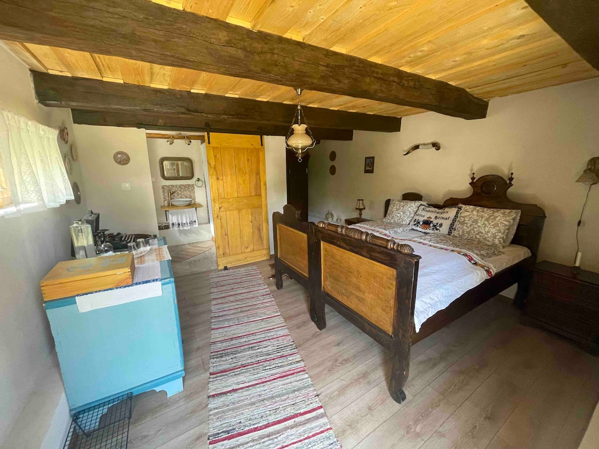A spacious bedroom features a wooden bed frame with decorative detail and crisp white linens. Natural light filters through a small window, and a colorful woven runner runs along the light-colored floor. A wooden door leads to another area, enhancing the room's rustic charm.