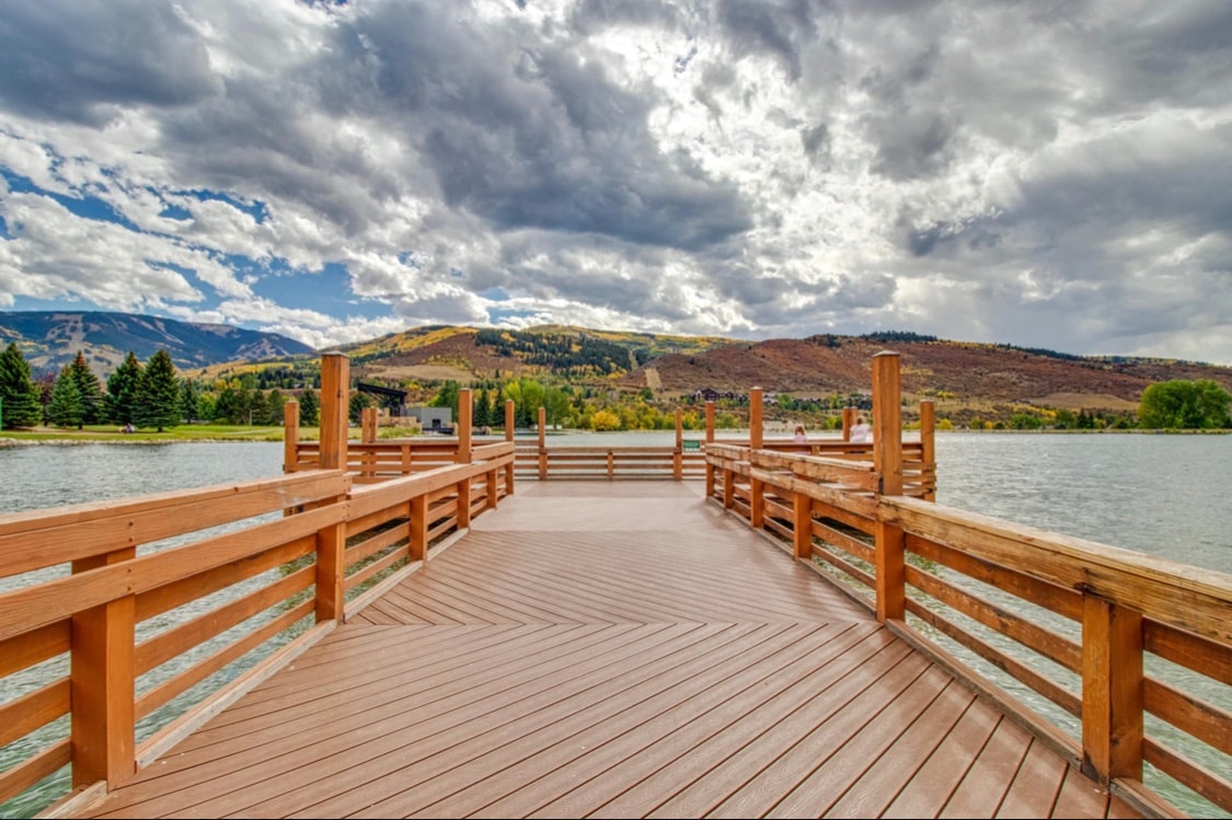 A wooden dock extends into a calm lake, surrounded by vibrant trees and rolling hills. Cloudy skies above contrast with the serene water, creating a peaceful outdoor atmosphere. The dock features sturdy railings, providing safety as one enjoys the scenic views of the landscape.