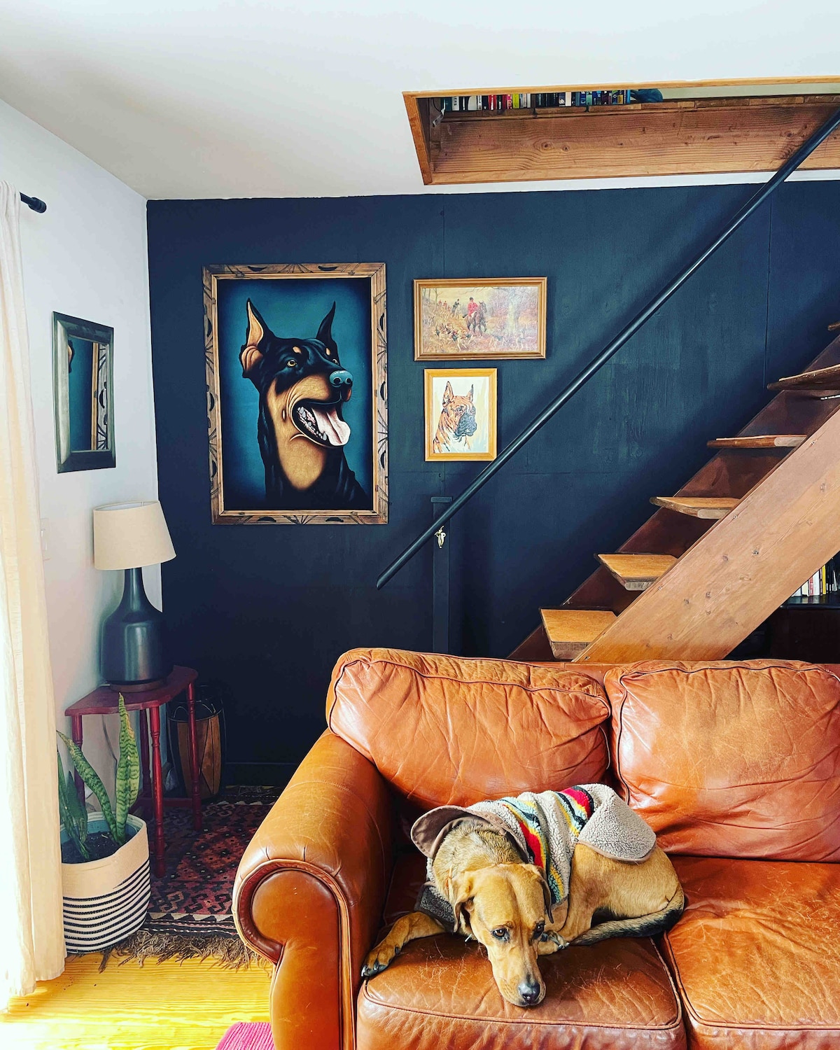 A cozy living room corner features a warm leather couch with a dog resting on it. A dark wall displays an artistic dog portrait, while wooden stairs lead to an upper level. A plant and lamp add subtle detail to the inviting space.
