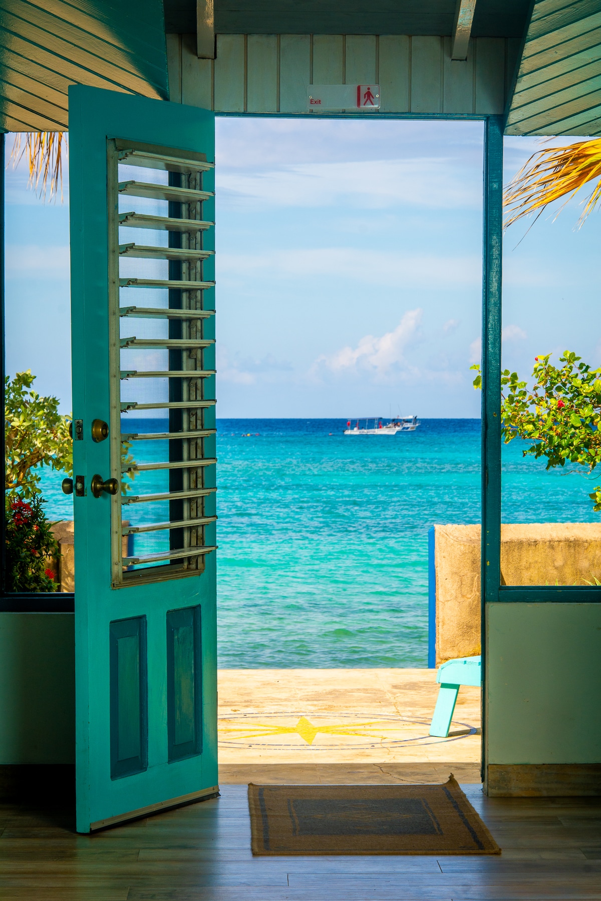 A bright blue door stands open, offering a view of the clear turquoise water and distant boat on the horizon. Lush greenery frames the entrance, enhancing the connection between the interior space and the inviting beach just outside.