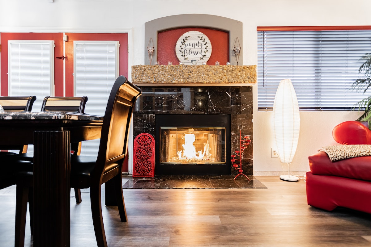 A welcoming living space features a modern fireplace with a stylish mantle. Nearby, a dining table is set against the backdrop of large windows, and a floor lamp provides ample lighting. Red accents bring warmth, complementing the neutral tones of the furniture.