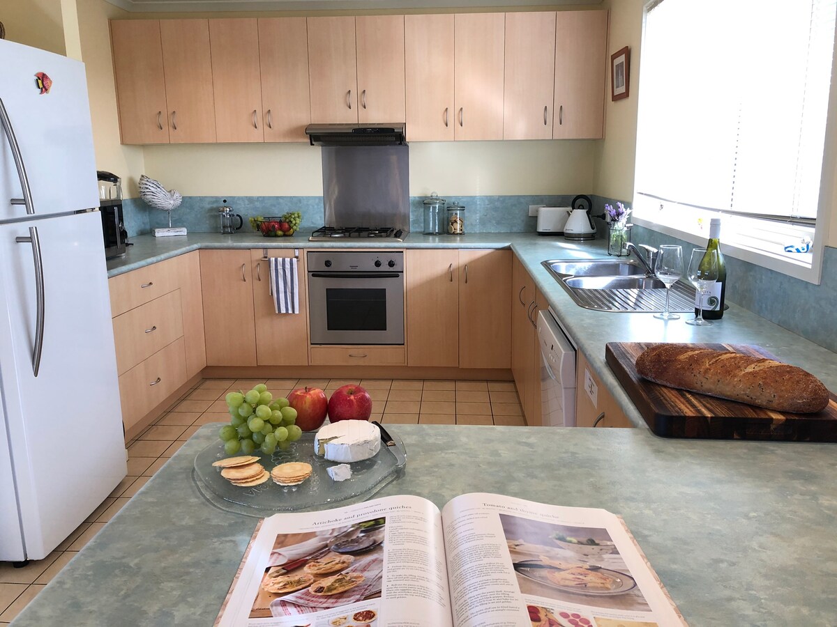 A functional kitchen displays light wood cabinetry and a teal countertop. A variety of fresh fruits, a loaf of bread, and cheese are arranged on a cutting board. A cookbook lies open on the counter, and sunlight illuminates the space through a window.