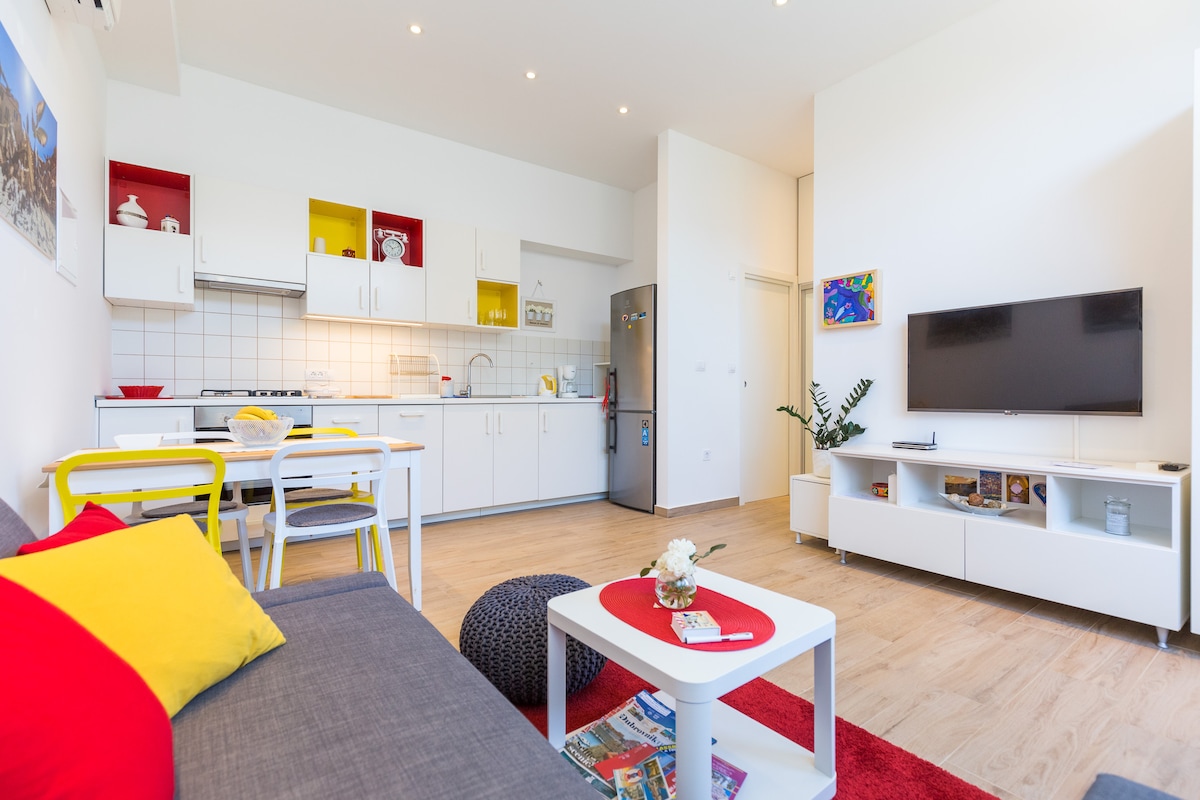 A modern kitchen and living area are depicted, featuring white cabinetry with red and yellow accents. A cozy grey sofa, adorned with bright cushions, faces a flat-screen TV. A dining table is set nearby, and light hardwood flooring adds warmth to the space.