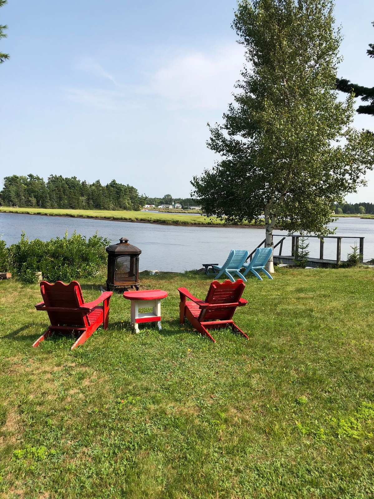 Adirondack chairs in red and blue are positioned on a grassy area by the Tidnish River, enhancing the outdoor seating space. A small table is placed between the chairs, and a lantern stands nearby. A view of the river and trees can be seen in the background.