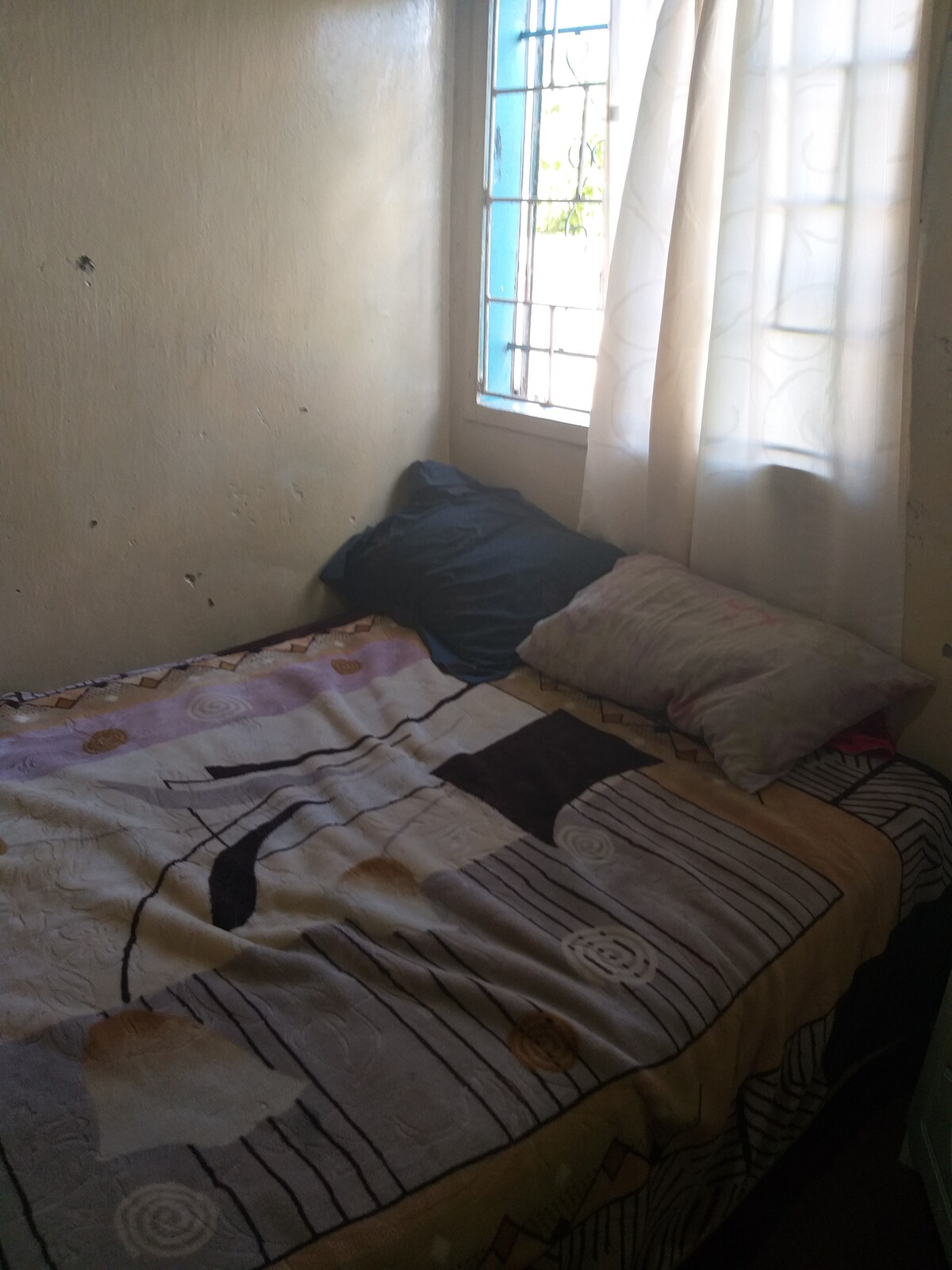 A compact sleeping area features a single bed adorned with a patterned blanket and decorative pillows. Natural light enters through a nearby window, partially dressed with sheer curtains, illuminating the warm wall color.