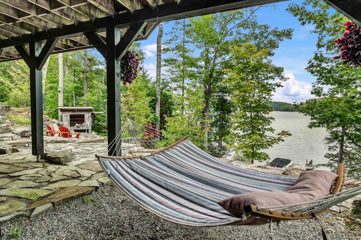 A serene outdoor space features a striped hammock nestled between trees, providing a tranquil spot for relaxation. Nearby, a stone patio leads to a fire pit area, accented by two red chairs. The gentle waters of the lake can be seen in the background.