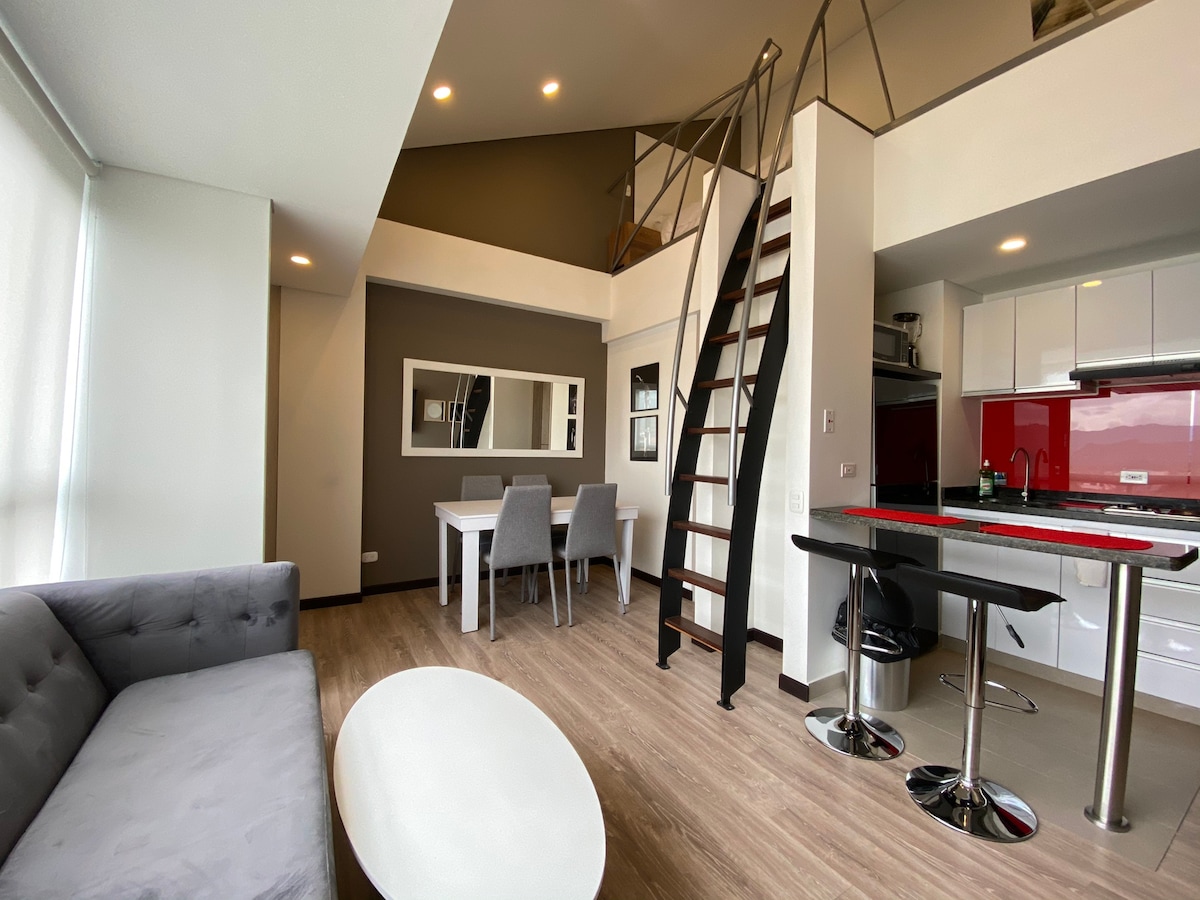 An open living space features a gray sofa and a circular coffee table, alongside a dining area with white chairs. The kitchenette includes sleek cabinets and a pop of color from the red backsplash. A wooden staircase leads to the upper level, enhancing the duplex design.