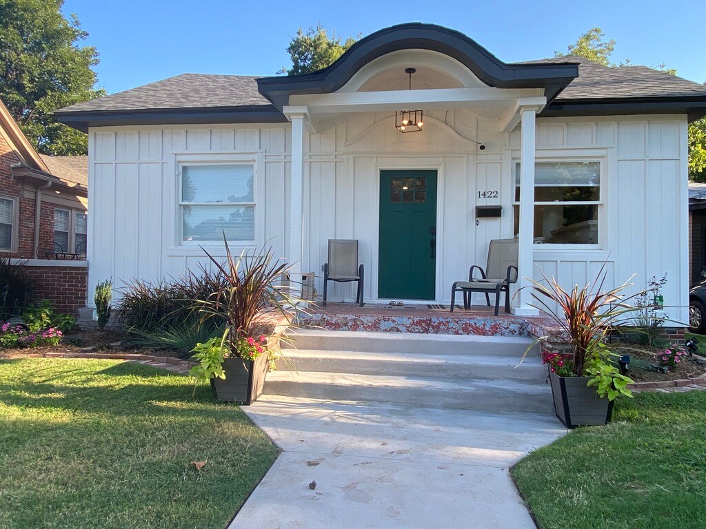 The front exterior of the cottage features a white façade with a green door, framed by two large windows. A small porch accommodates two chairs, and colorful flower pots enhance the entrance. A well-maintained lawn leads up to the concrete pathway.