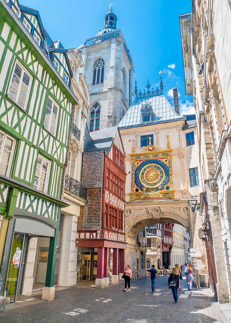 A charming street scene showcases historic architecture with intricately detailed facades in varied colors. A large ornate clock hangs above an archway, capturing attention. People can be seen walking leisurely beneath a bright blue sky, highlighting the vibrant atmosphere of the area.