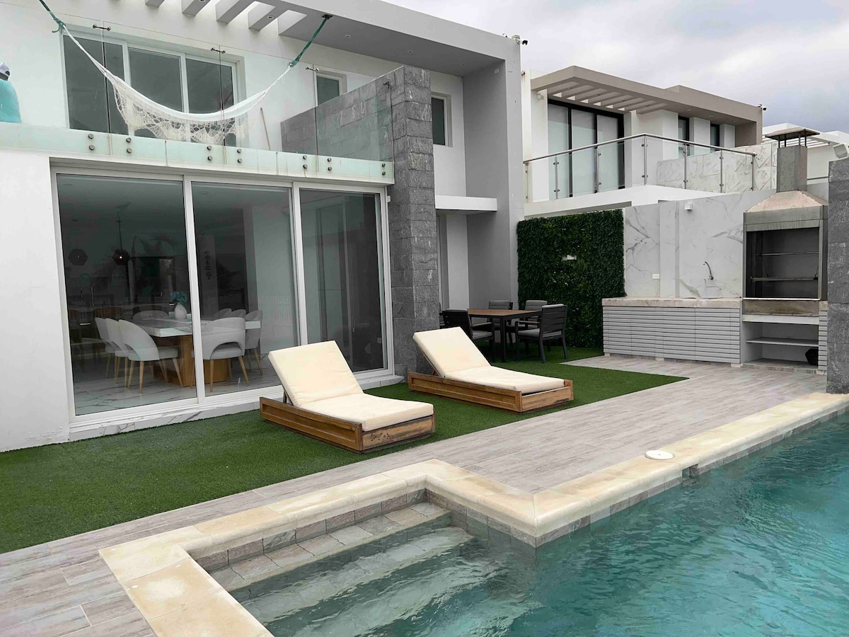 An outdoor area is presented with two lounge chairs positioned beside a turquoise pool. A dining table is visible in the background, surrounded by greenery. The modern architecture of the house features large glass doors, allowing natural light to fill the space.