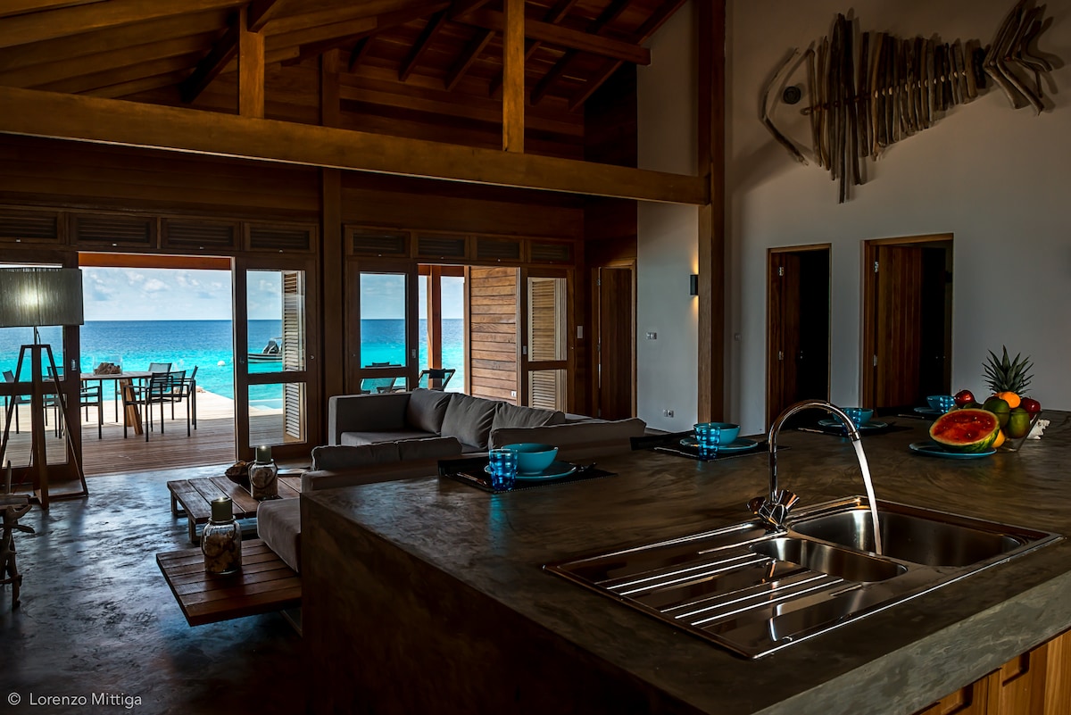 The open-plan living area features a spacious kitchen with a modern sink and countertop. Soft seating arranged with a view of the ocean enhances the inviting atmosphere. Large glass doors provide a seamless connection to the outdoor space, while natural light illuminates the interior.
