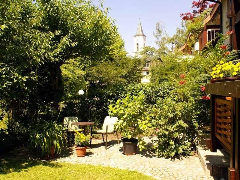 A tranquil garden space features lush greenery and potted plants. A small table and two chairs set among the foliage invite relaxation. In the background, a church tower is visible, framed by trees, creating a serene outdoor atmosphere.