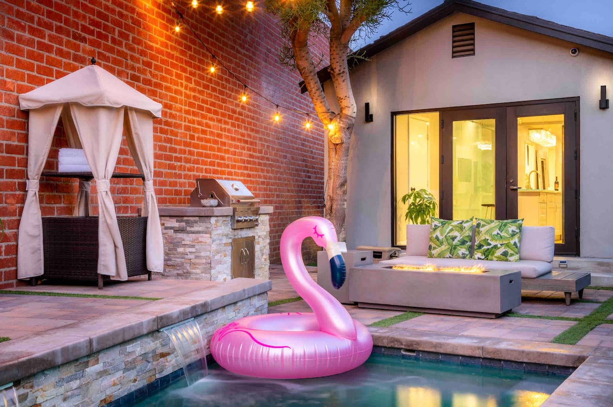 An outdoor space features a small dipping pool with a pink flamingo float. A cozy seating area with a firepit is surrounded by pavers. An outdoor kitchen is seen next to a shaded gazebo. String lights cast a soft glow across the area as evening approaches.