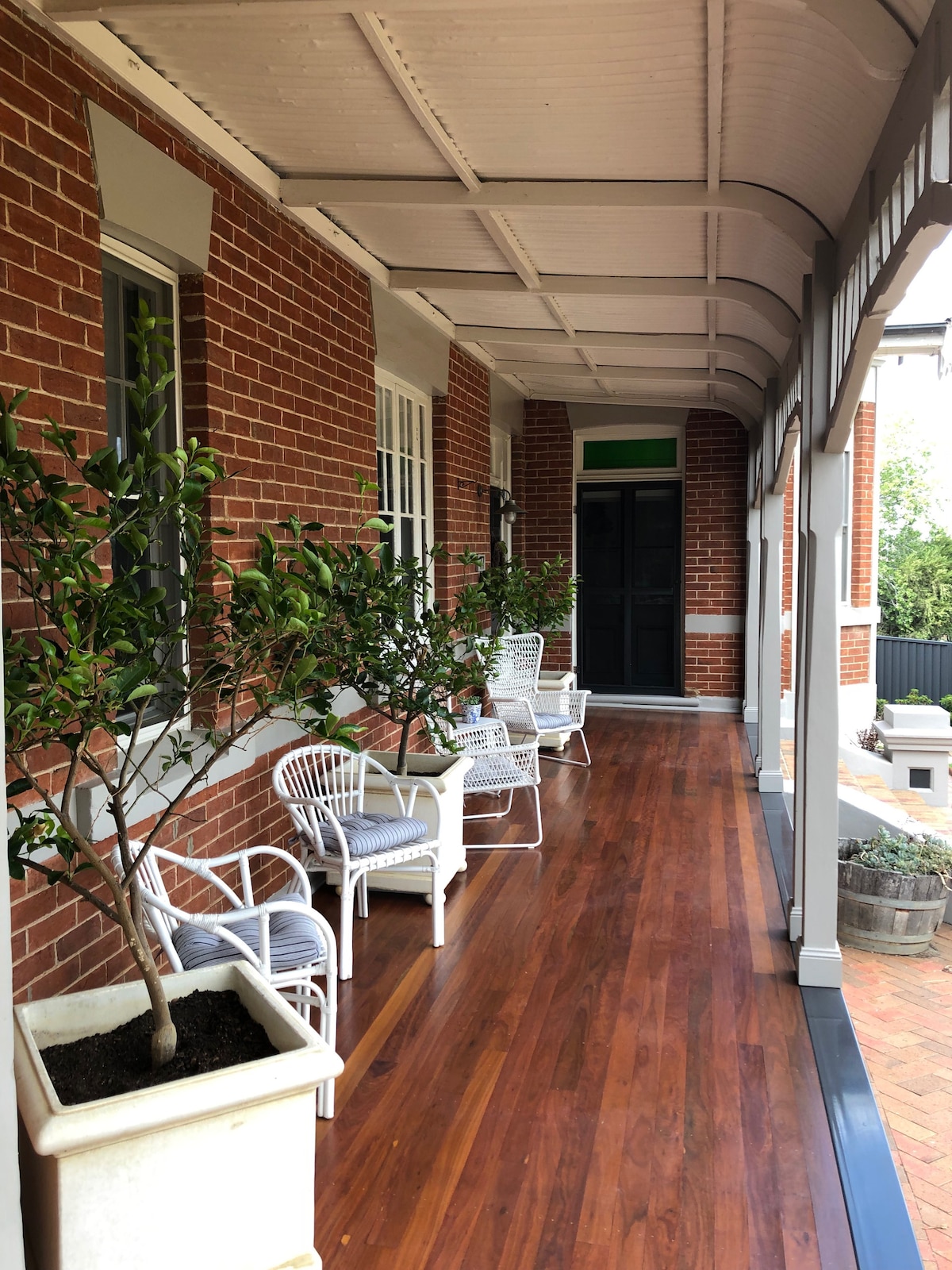 A covered porch features several white chairs arranged alongside potted plants. The warm wooden flooring contrasts with the red brick walls of the home. A black front door is visible in the background, framed by large windows that enhance the open feel.