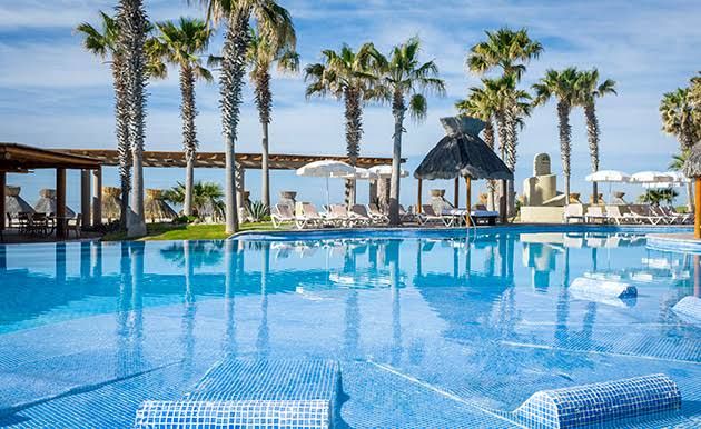 A tranquil swimming pool features crystal-clear water surrounded by palm trees. Comfortable lounge chairs and umbrellas are positioned within a relaxing outdoor area, offering shade and leisure. An open structure with a thatched roof provides additional seating space nearby.