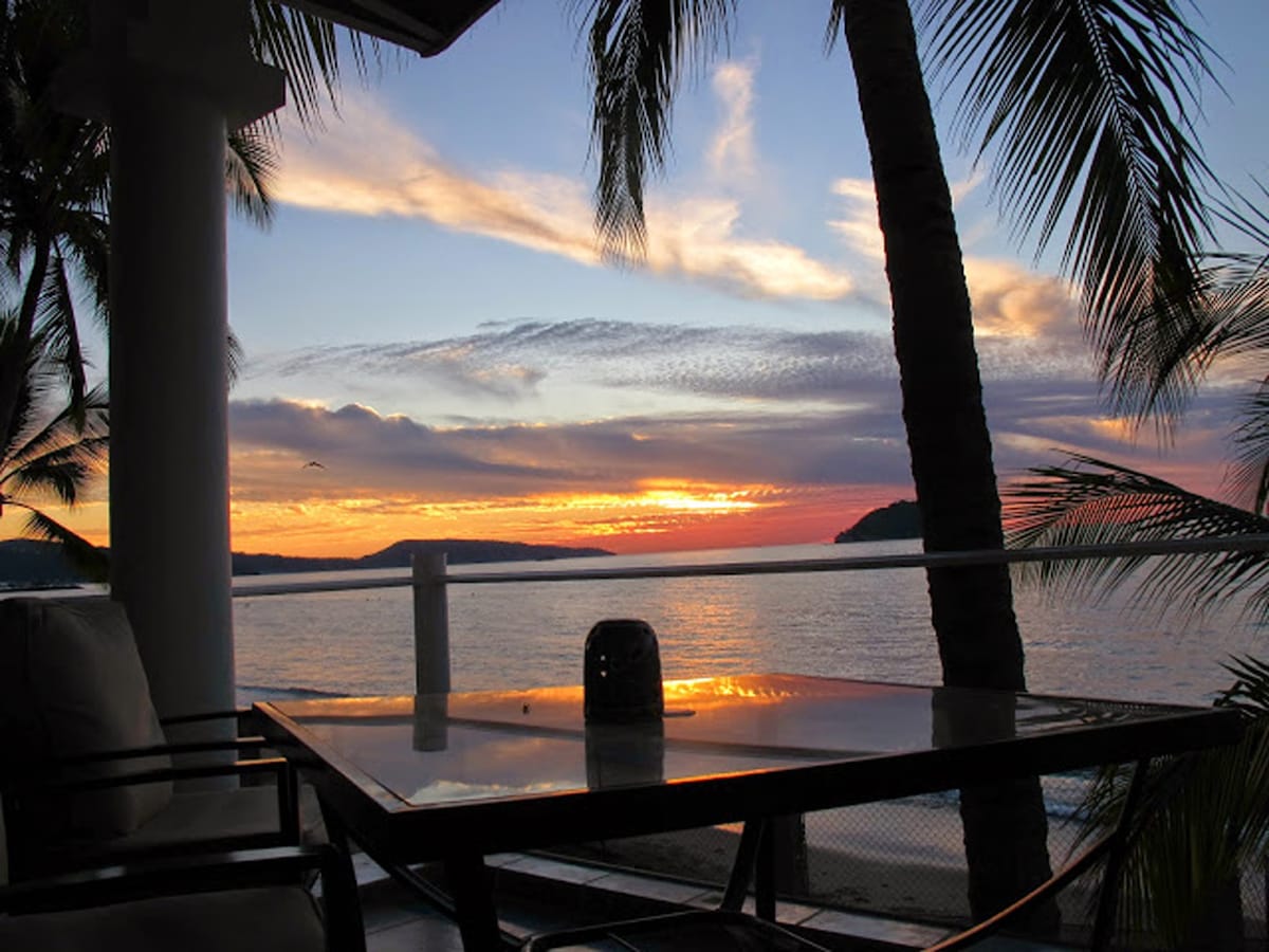 A serene sunset view is captured from an outdoor deck, featuring a glass table surrounded by black chairs. Palm trees frame the scene, and the vibrant colors of the sky reflect on the calm ocean waters, enhancing the peaceful ambiance of the beachfront location.