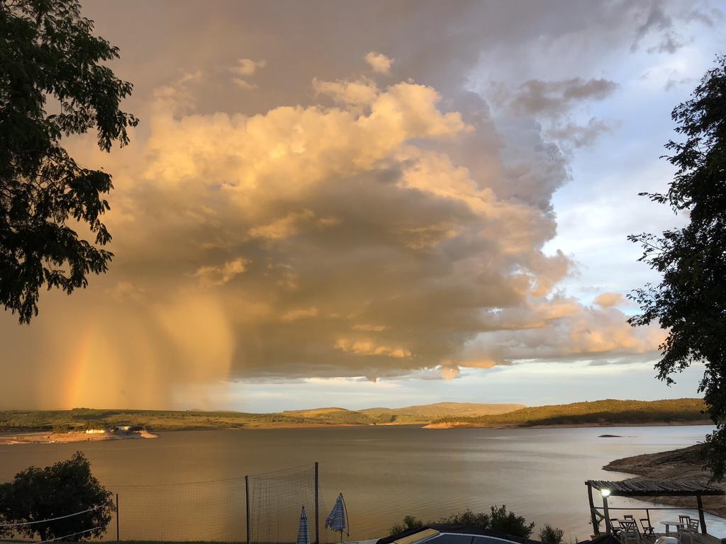 A serene lakeside view showcases a tranquil body of water bordered by green hills. Prominent clouds in various shades of gold and gray fill the sky, while a faint rainbow can be seen in the distance. Sunlight reflects gently on the water's surface.
