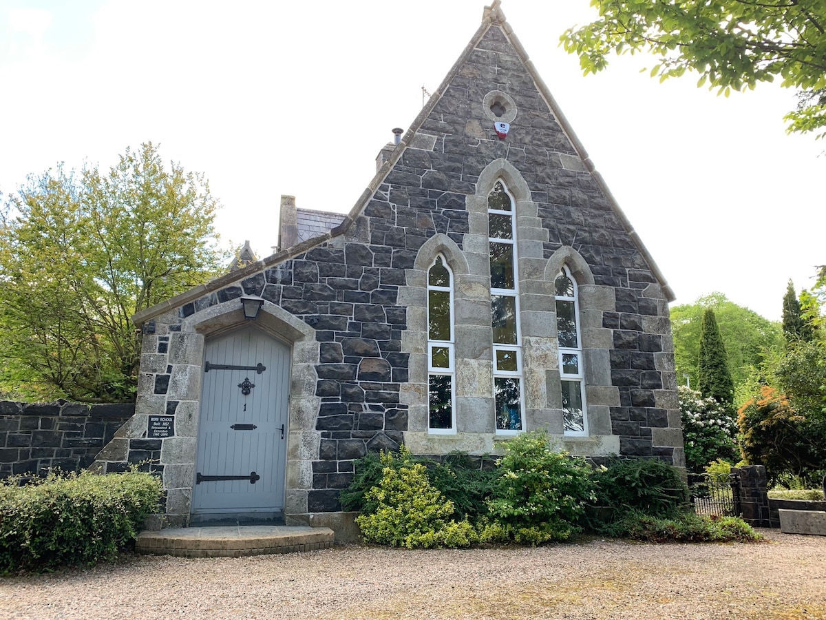 The exterior of a historic stone building is shown, featuring tall, arched windows with clear glass. A decorative wooden door lies at the center, flanked by lush greenery and well-maintained shrubs. The structure is set against a bright sky, enhancing its elegant architecture.