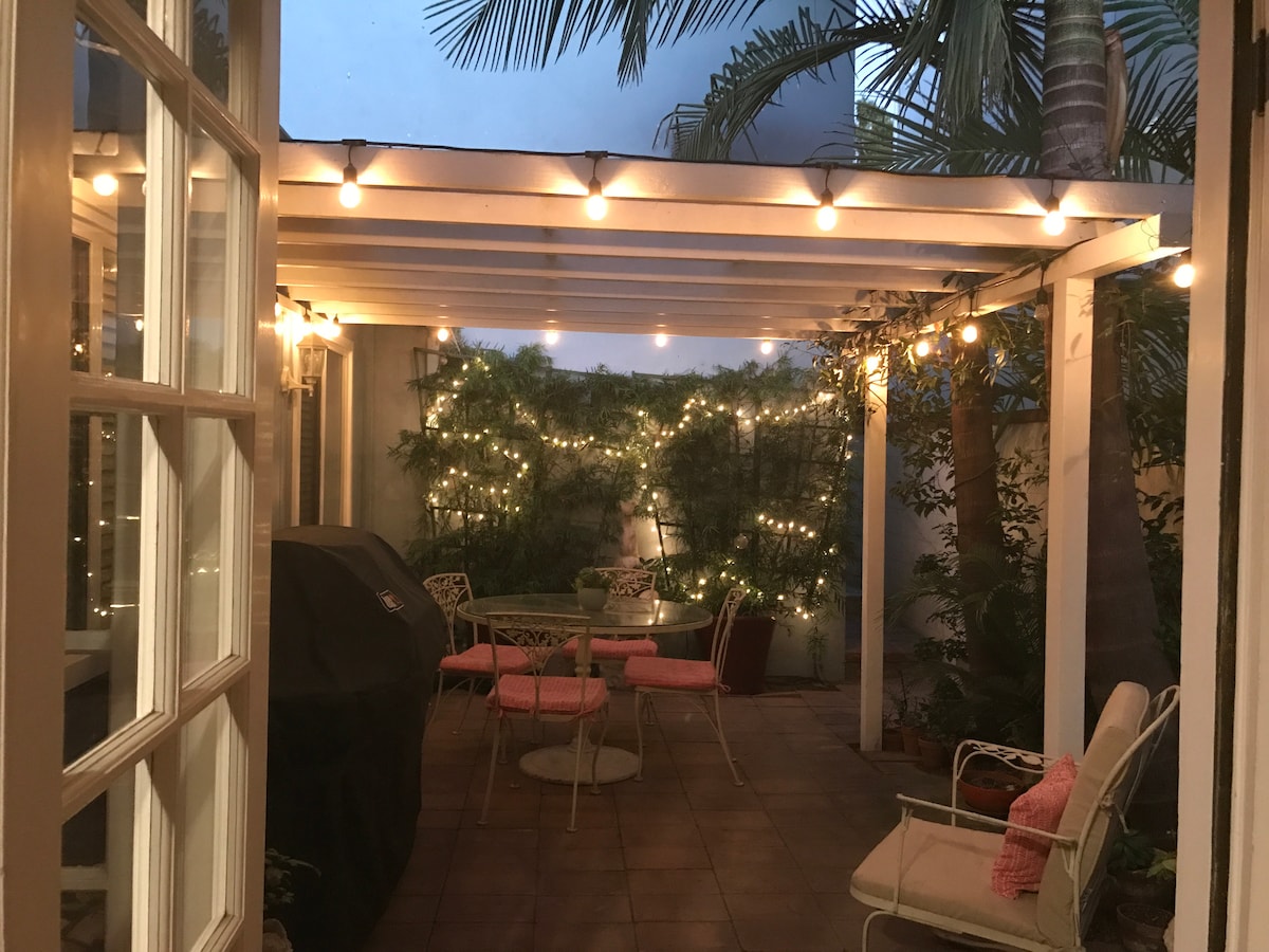An outdoor courtyard is depicted, featuring a covered seating area with a round table and four chairs. Soft lighting is provided by string lights above, and lush greenery surrounds the space, creating a serene atmosphere. A Weber gas BBQ is visible in the corner.