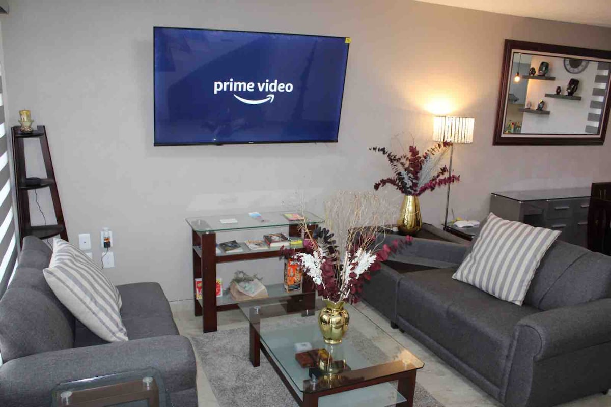 A comfortable living area is equipped with a wall-mounted television displaying Prime Video. Two plush sofas are positioned around a glass coffee table, complemented by decorative elements like a vase filled with dried flowers. Soft lighting enhances the cozy ambiance.