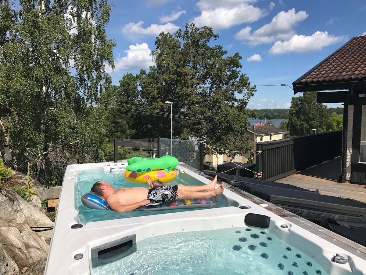 A swim spa is featured on a sunlit terrace, surrounded by greenery and offering views of the lake. An individual relaxes on a float, showcasing the spacious area designed for leisure and enjoyment. Nearby, the deck offers ample space for outdoor seating and social activities.