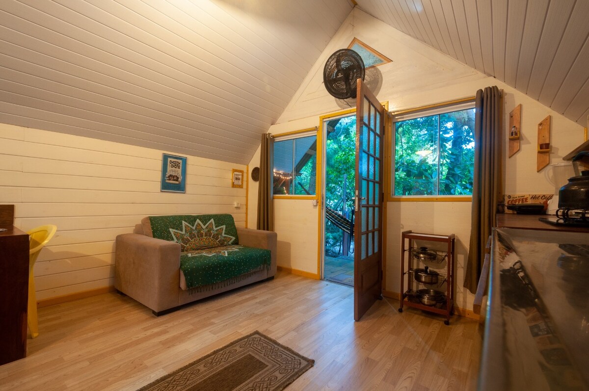 A bright living area features a soft couch adorned with intricate patterns, placed against a wall of white wooden panels. Large windows allow natural light to fill the space, offering views of the surrounding greenery. A small dining area is visible, enhancing the cozy ambiance.
