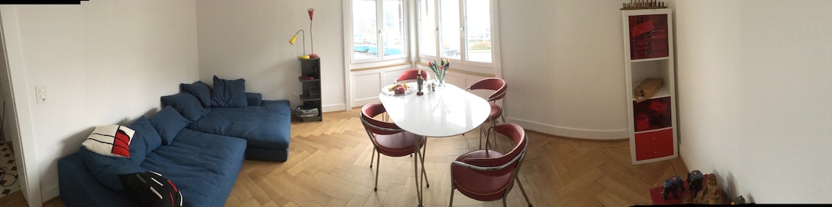 A spacious dining area features a round white table surrounded by six red chairs. A blue sofa is positioned nearby, and a vase of flowers adds a colorful touch. Natural light fills the room through large windows, highlighting the wooden flooring.