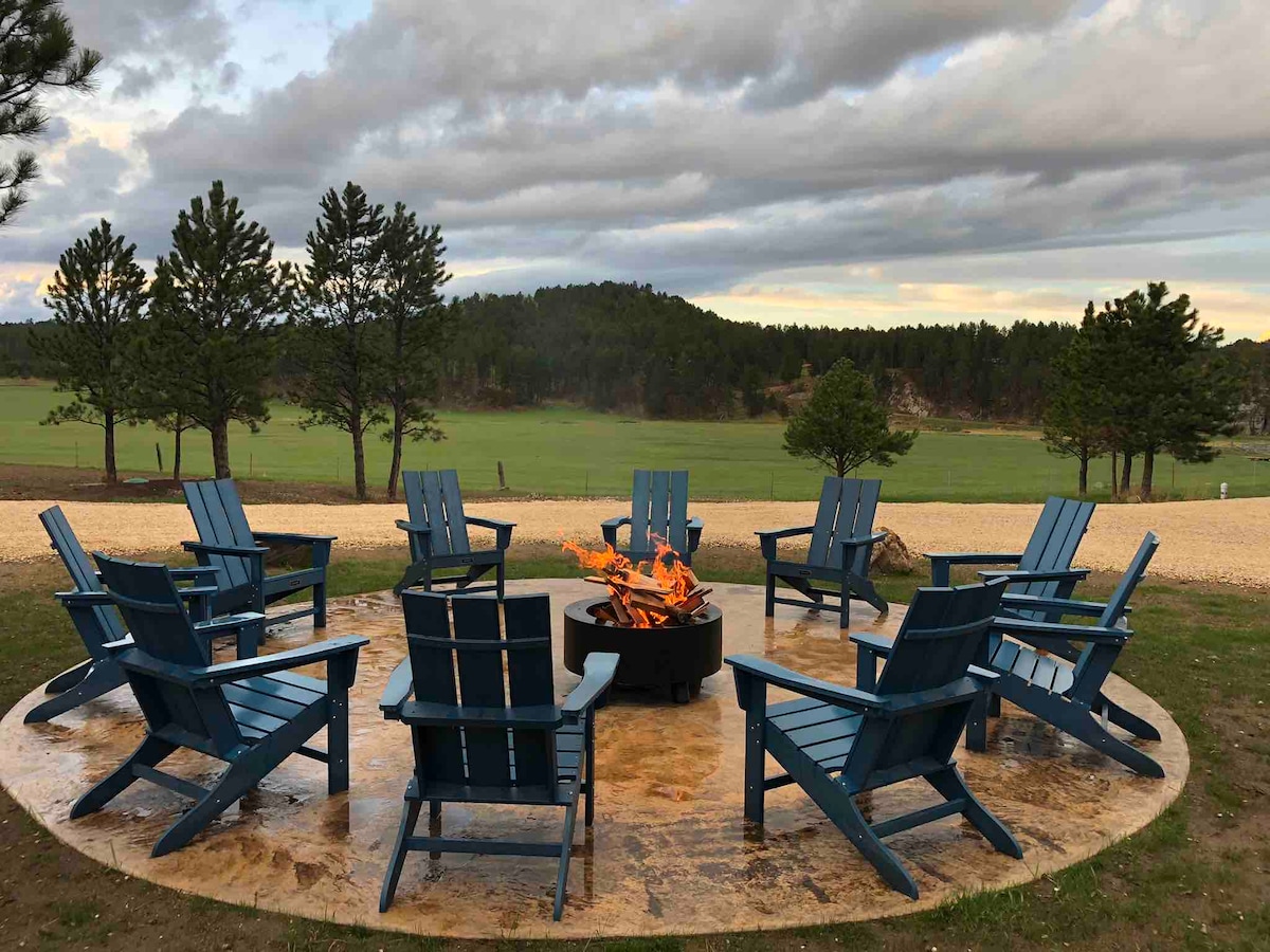 A circular seating area features ten blue Adirondack chairs arranged around a central fire pit. Natural landscaping and trees surround the space, with open grassy fields extending into the background. The sky displays a blend of clouds and soft light, suggesting an inviting atmosphere for gathering.