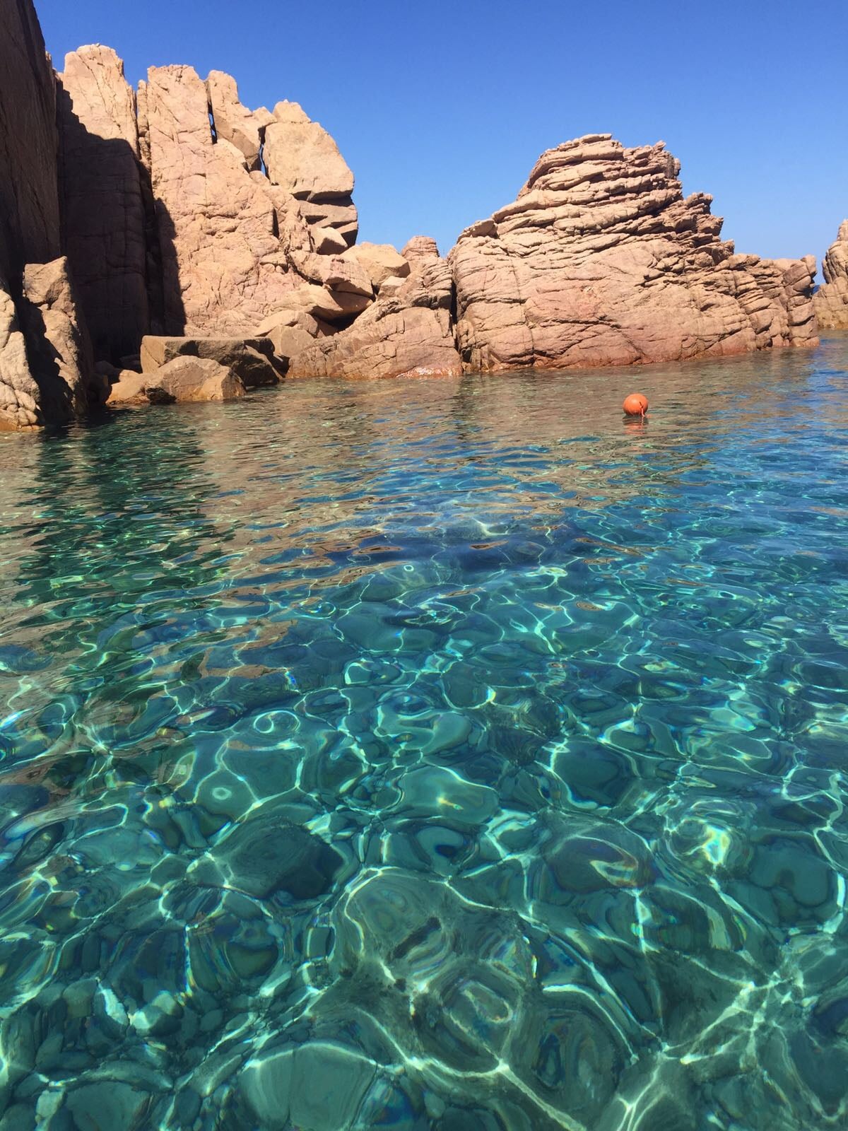 A clear, vibrant blue water is seen, reflecting the sunlight with a shimmering effect. Rocky formations rise from the water's edge, adding natural texture to the scene. The clarity of the water reveals a sandy bottom, creating an inviting atmosphere for swimming.
