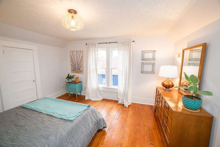 Upstairs master bedroom, with a queen bed,  vintage full-size dresser, and large closet. Views out the window show you the blues, greens, and lemon yellow of surrounding homes and churches.