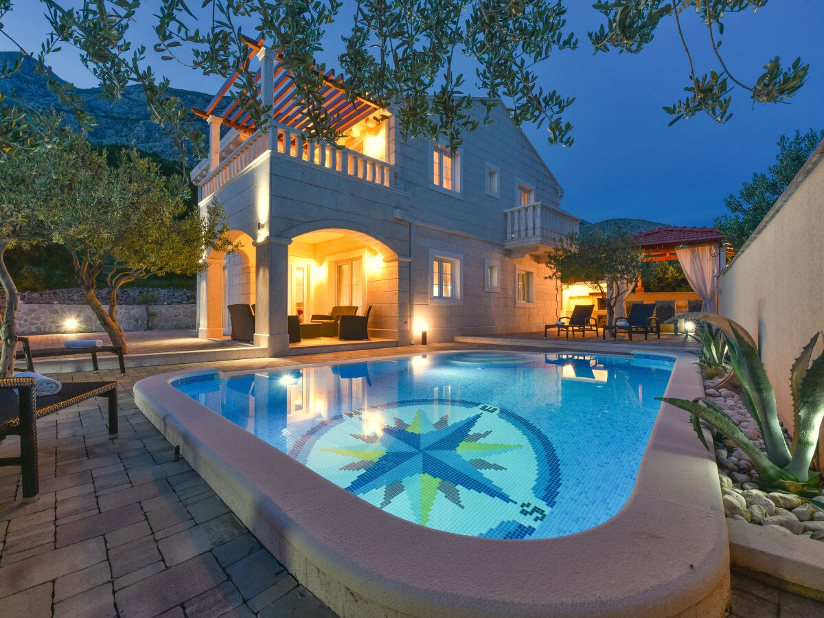 A modern villa is showcased at dusk, featuring a private pool with a compass mosaic design. Surrounding palm and olive trees create a lush environment, while outdoor seating areas are positioned near the pool. Soft lighting highlights the villa's stone façade and arched balcony.