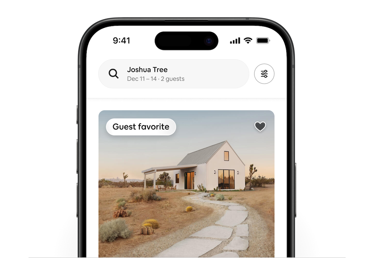 A phone shows an Airbnb listing of a white farm house in Joshua Tree. In the top-left corner of the listing is a Guest Favorite badge.