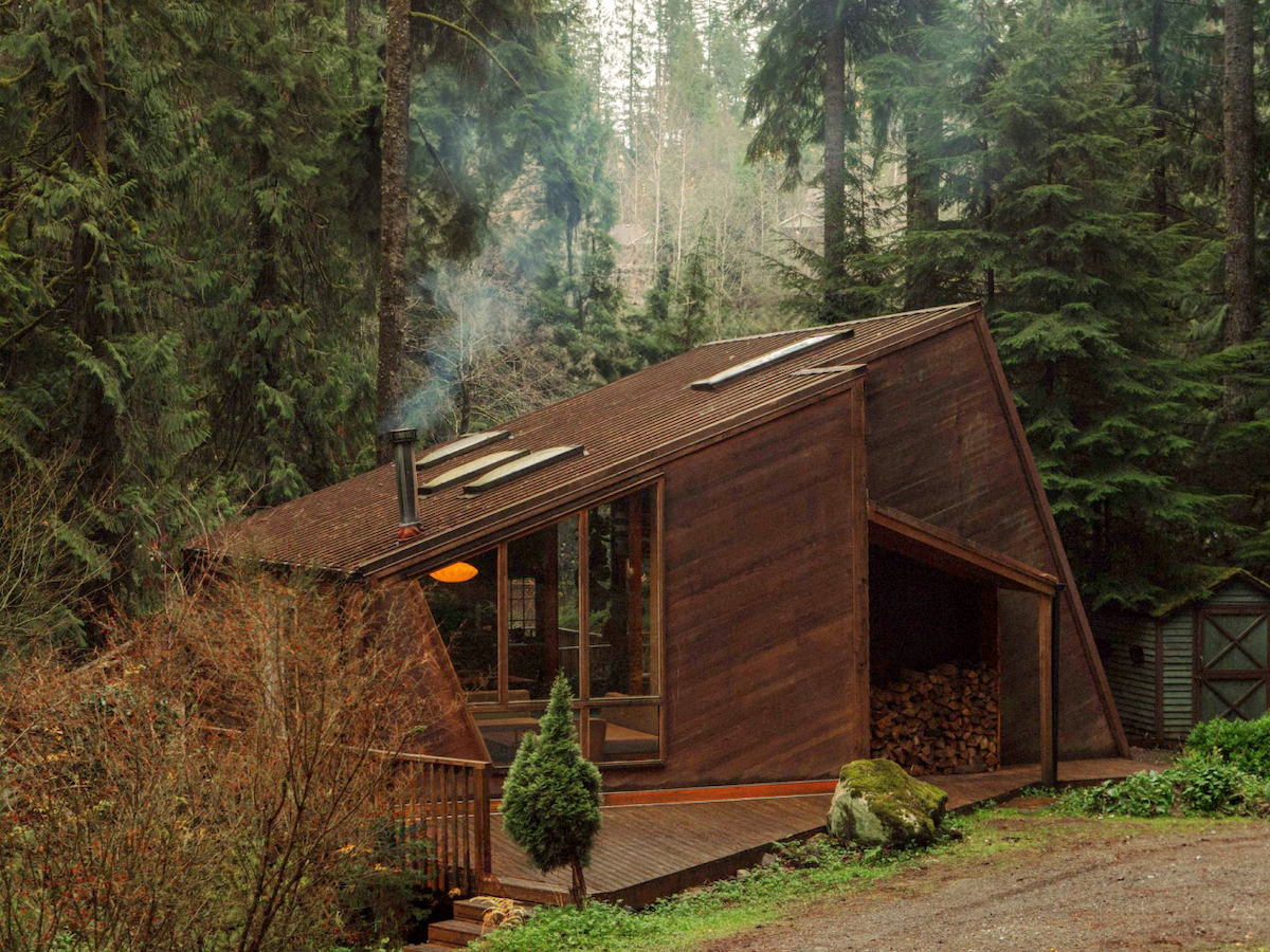 A brown cabin with a slanted roof surrounded by tall evergreens in a dense forest. Smoke rises from the chimney.