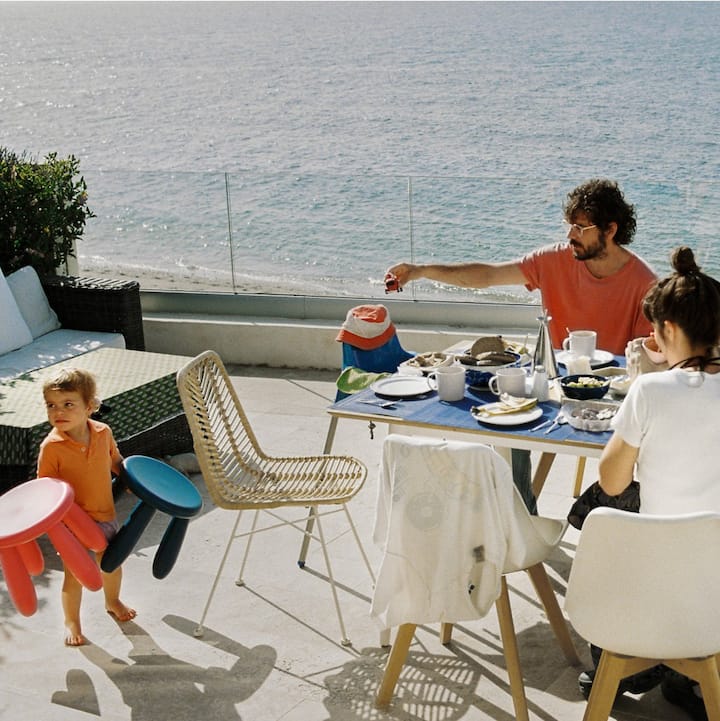 Two men, a woman, and a toddler at an outdoor table overlooking a beach and water. 