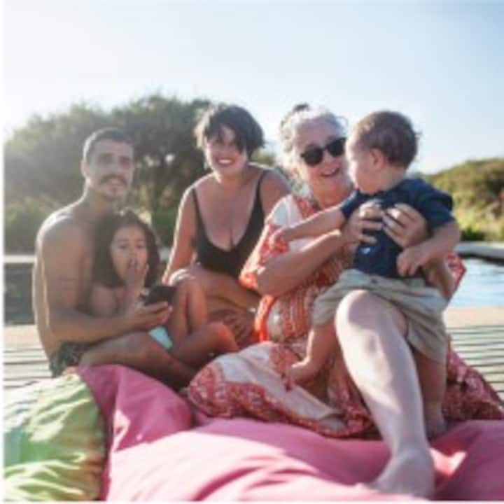 Multi-generational family with two young kids, parents, and a grandma sitting in the sun next to a pool.