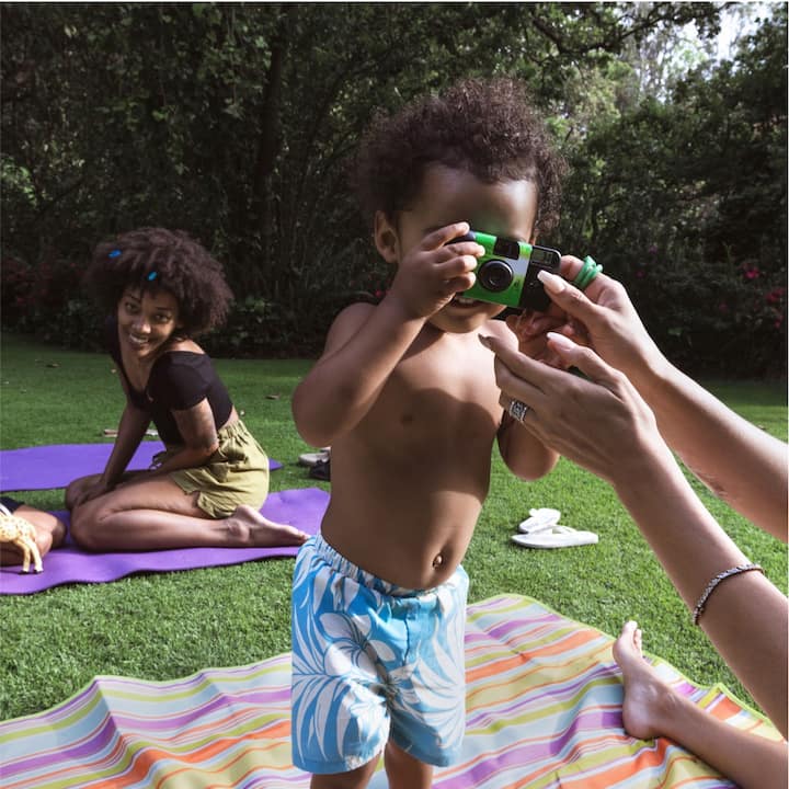 A young woman and two young children outdoors on yoga mats. One child is holding a camera and pointing it at the viewer. 