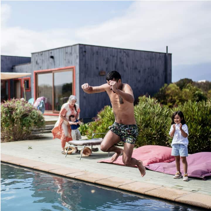A man jumping into a pool in front of a modern, box-style house while two children and a woman look on. 