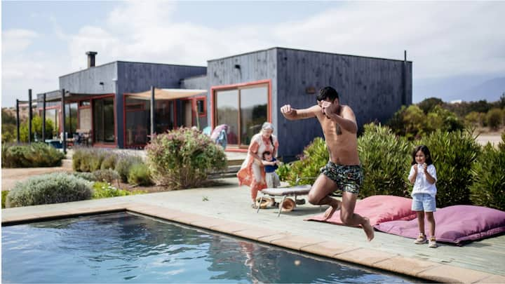 A man jumping into a pool in front of a modern, box-style house while two children and a woman look on. 