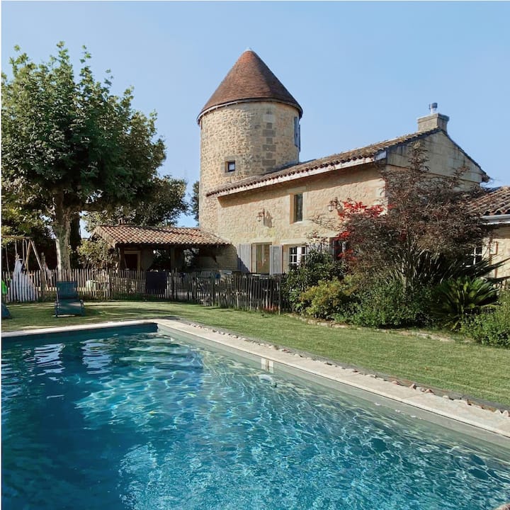 A swimming pool in the lawn outside of an old stone farmhouse with a tile roof and turret. 