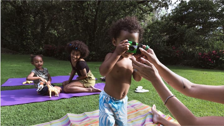 A young woman and two young children outdoors on yoga mats. One child is holding a camera and pointing it at the viewer. 