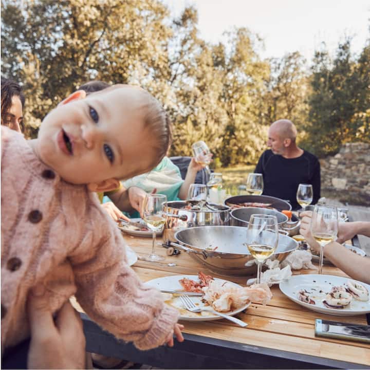A family eating a meal at an outdoor table with the baby in the foreground looking directly at the camera. 
