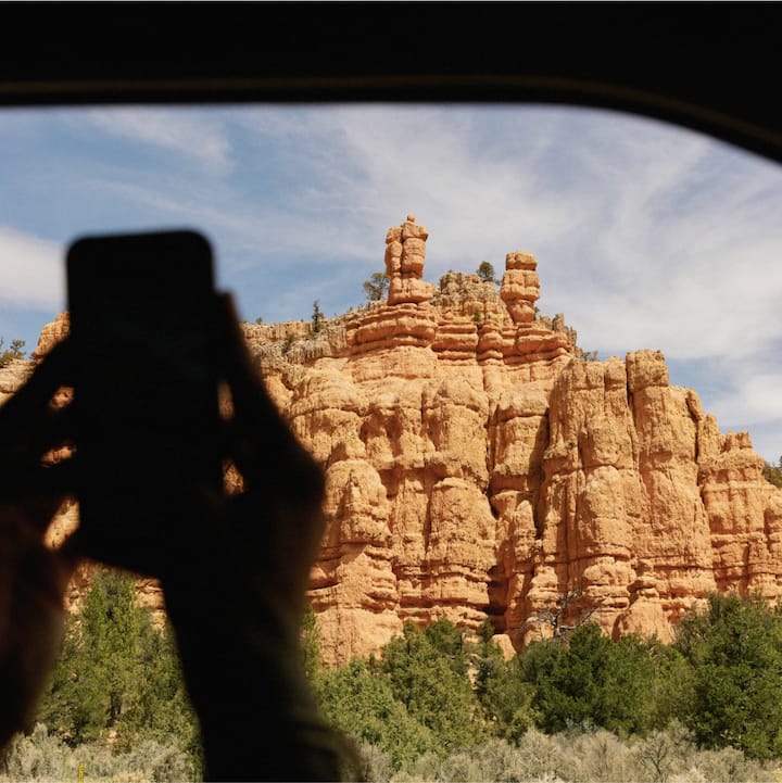 Backlit image of a person holding a smartphone taking a photo of some rocky hills out of a car window. 